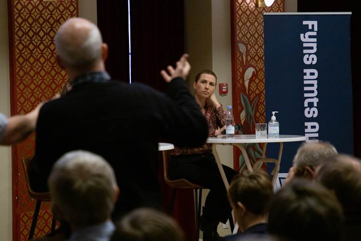 Mette Frederiksen sidder og lytter til et spørgsmål fra en avislæser. En mand står med en mikrofon i hånden 