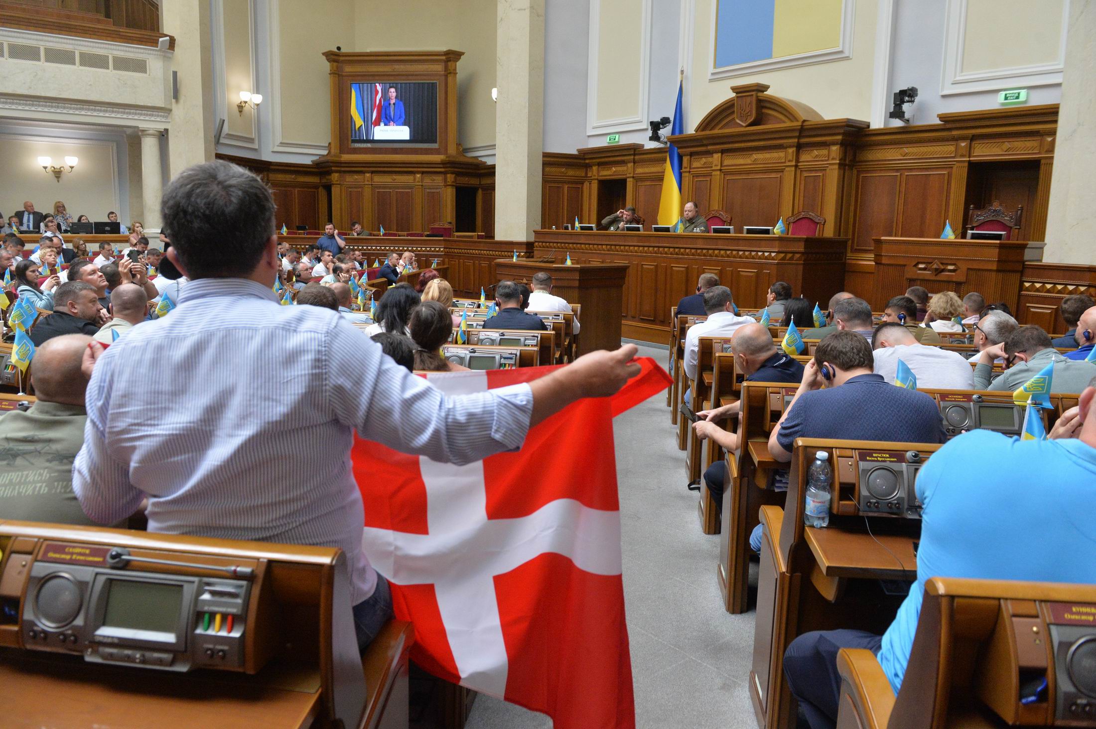 Statsminister Mette Frederiksen ses på storskærm tale til det ukrainske parlament, mens en tilhører holder Dannebrog.
