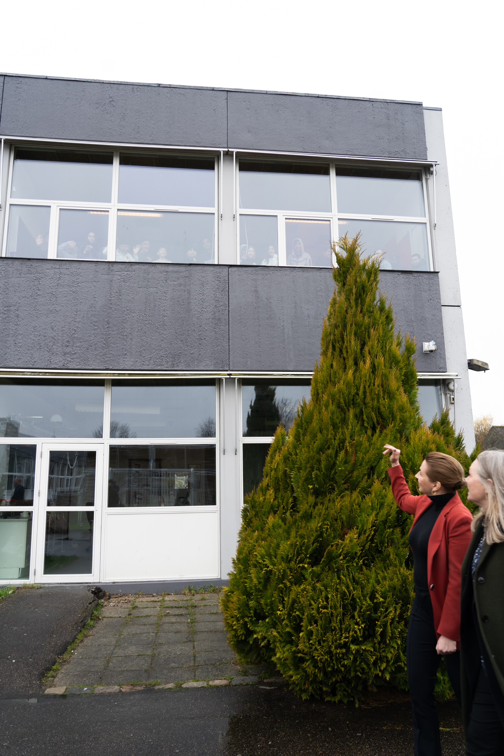 Statsminister Mette Frederiksen vinker til elever på skolen fra indgangen. Eleverne sidder på 1. sal. 