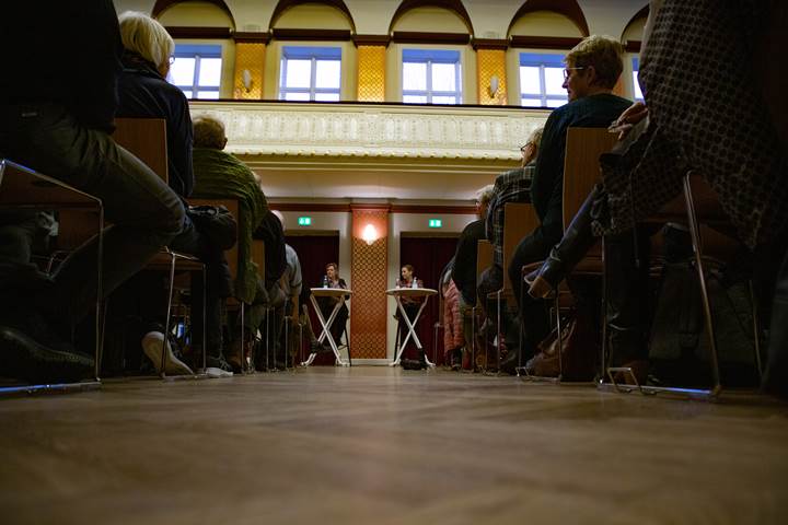 En gang mellem rækker af avislæsere, der er fremmødt til debat på Fyns Amts avis, ses nedefra gulvet. I enden står to rundeborde, hvor Statsminister Mette Frederiksen står ved det ene. 